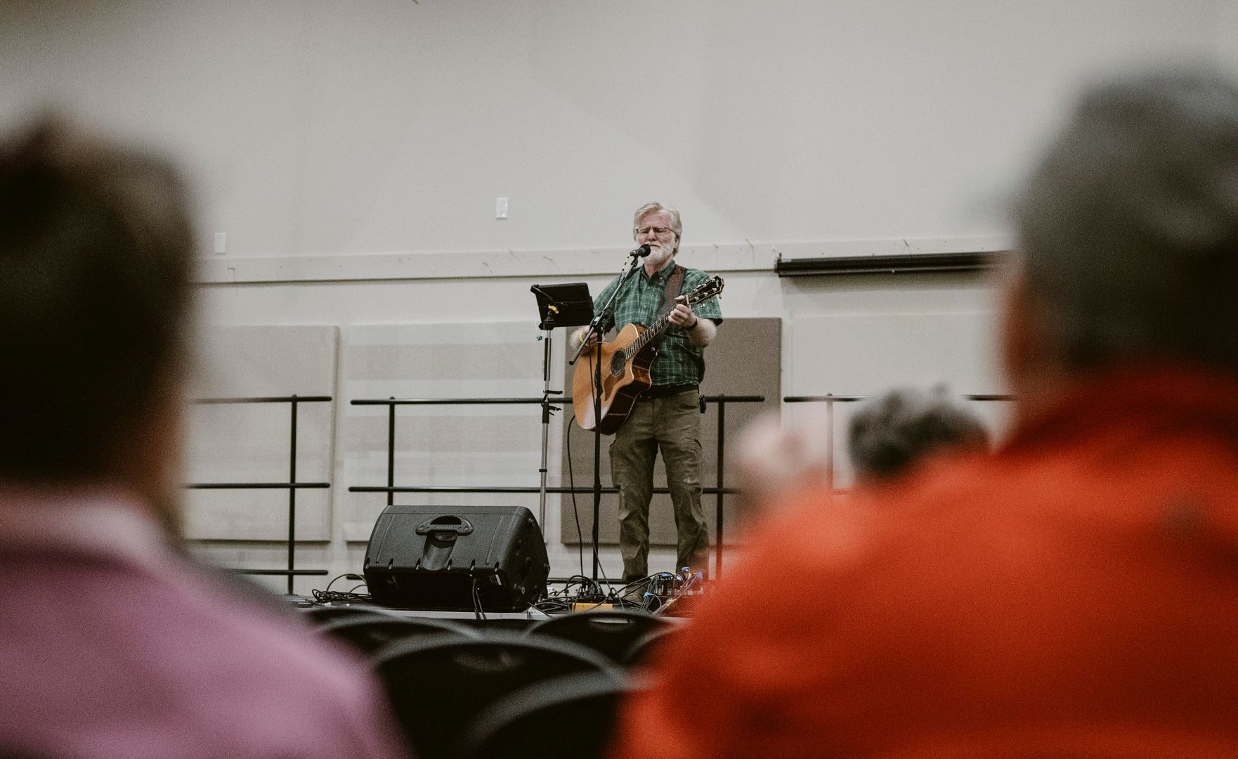 A musician plays an acoustic guitar and sings into a microphone on a stage in front of an audience.