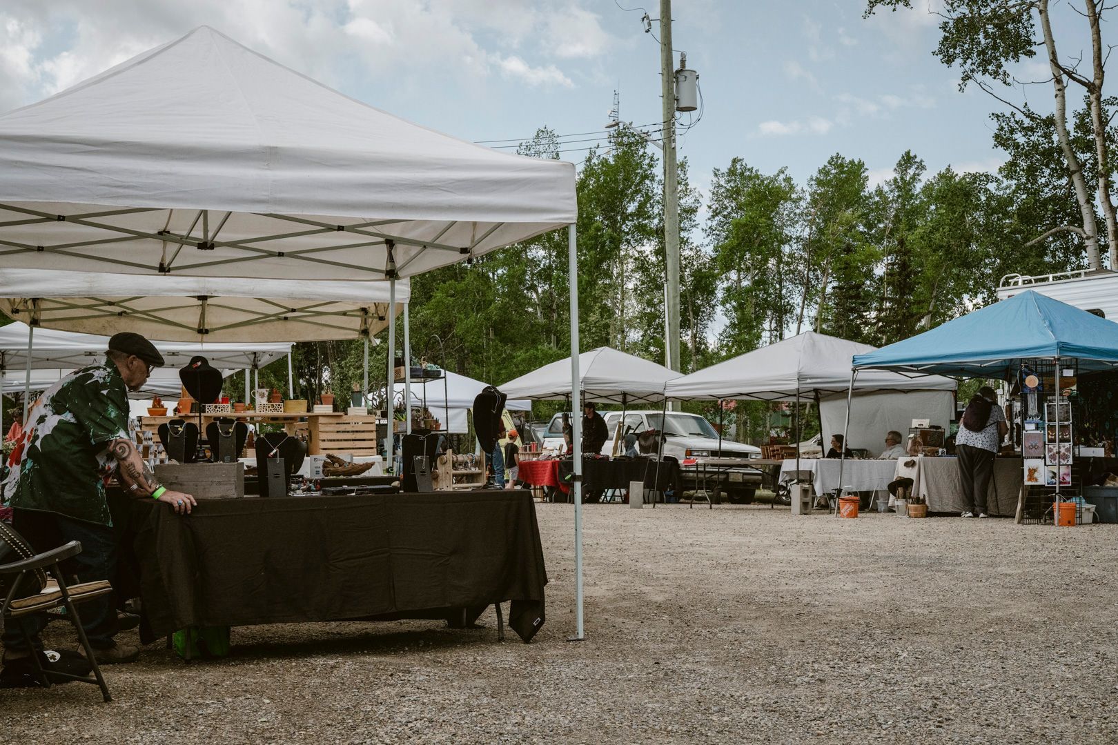 People browsing booths at an outdoor farmers market set up with white and blue tents on a gravel lot under a sunny sky.