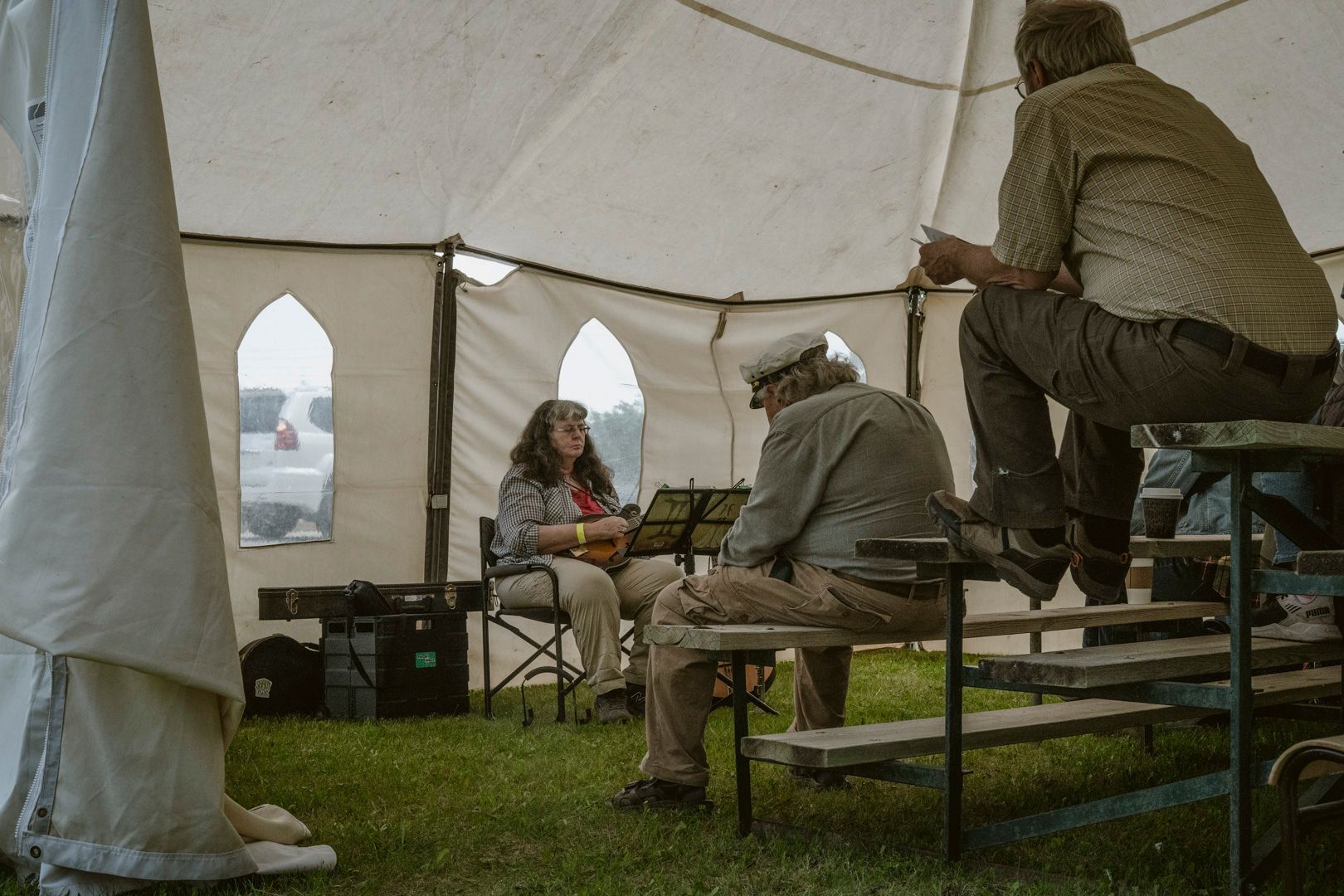 Two people seated on benches and a woman in a folding chair play musical instruments inside a large, light-colored tent.