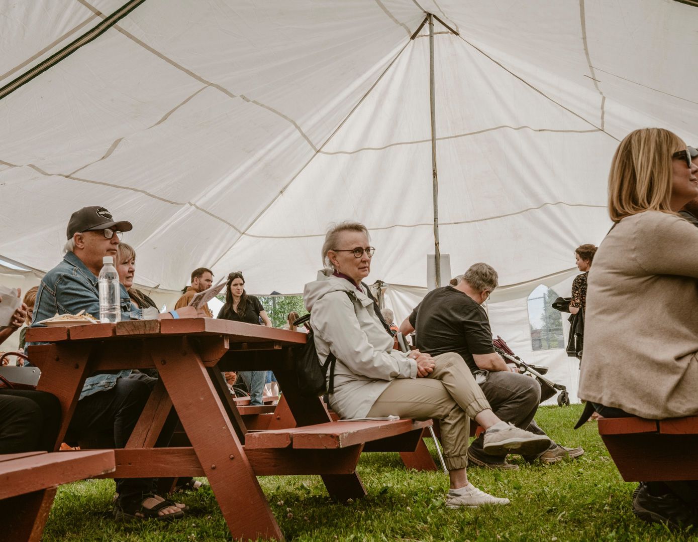 People sit at red picnic tables under a white canopy tent outdoors on a grassy lawn.