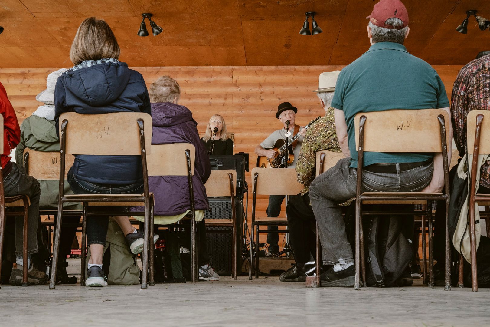 An audience sits on wooden chairs facing a performer with a guitar on a rustic wooden stage.