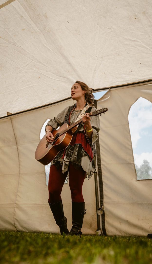 A performer plays an acoustic guitar inside a white tent, wearing a patterned top, red leggings, and dark boots.