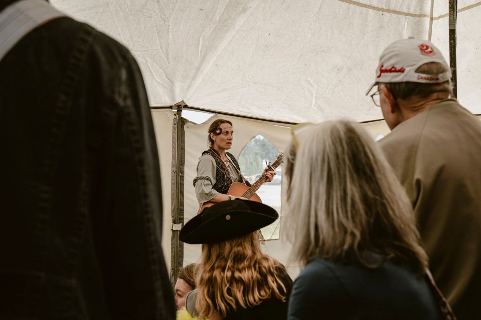 A musician performs with an acoustic guitar inside a tent while an audience watches from behind.