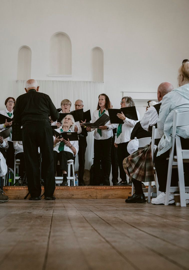 A conductor leads a small choir singing from binders on a wooden stage in a room with arched wall cutouts.