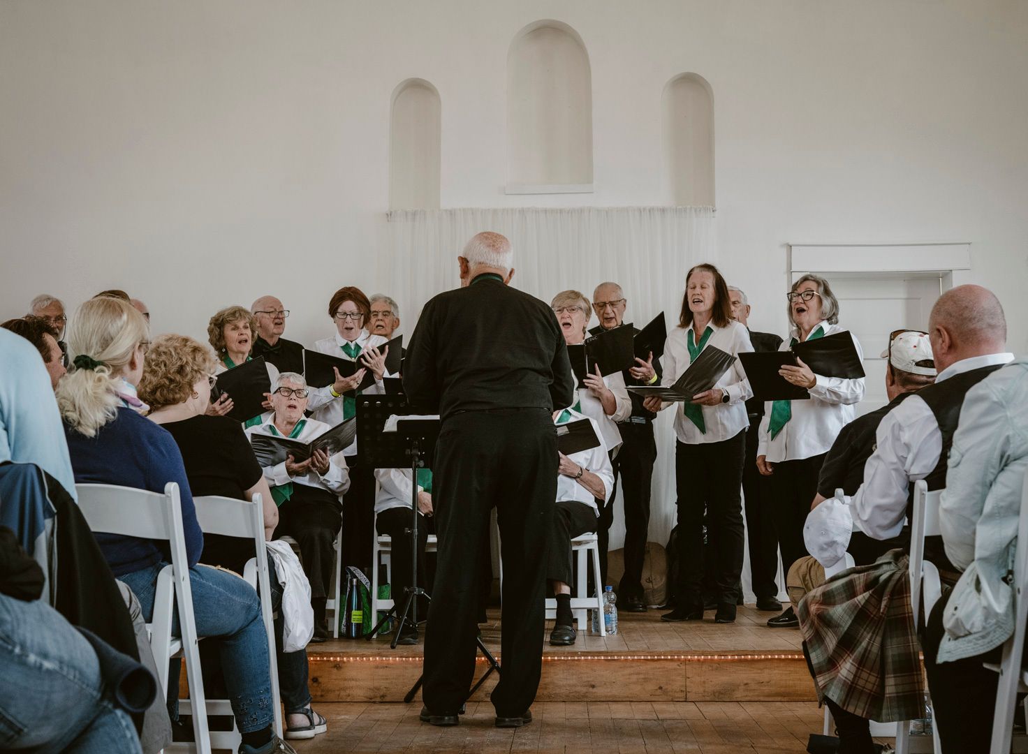 A choir performs on a stage in a simple room, led by a conductor standing with their back to the camera.
