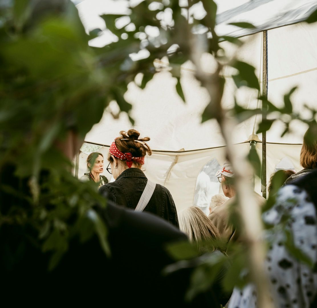 A person wearing a red bandana and dark clothing stands in a crowd beneath a white tent, framed by foreground leaves.