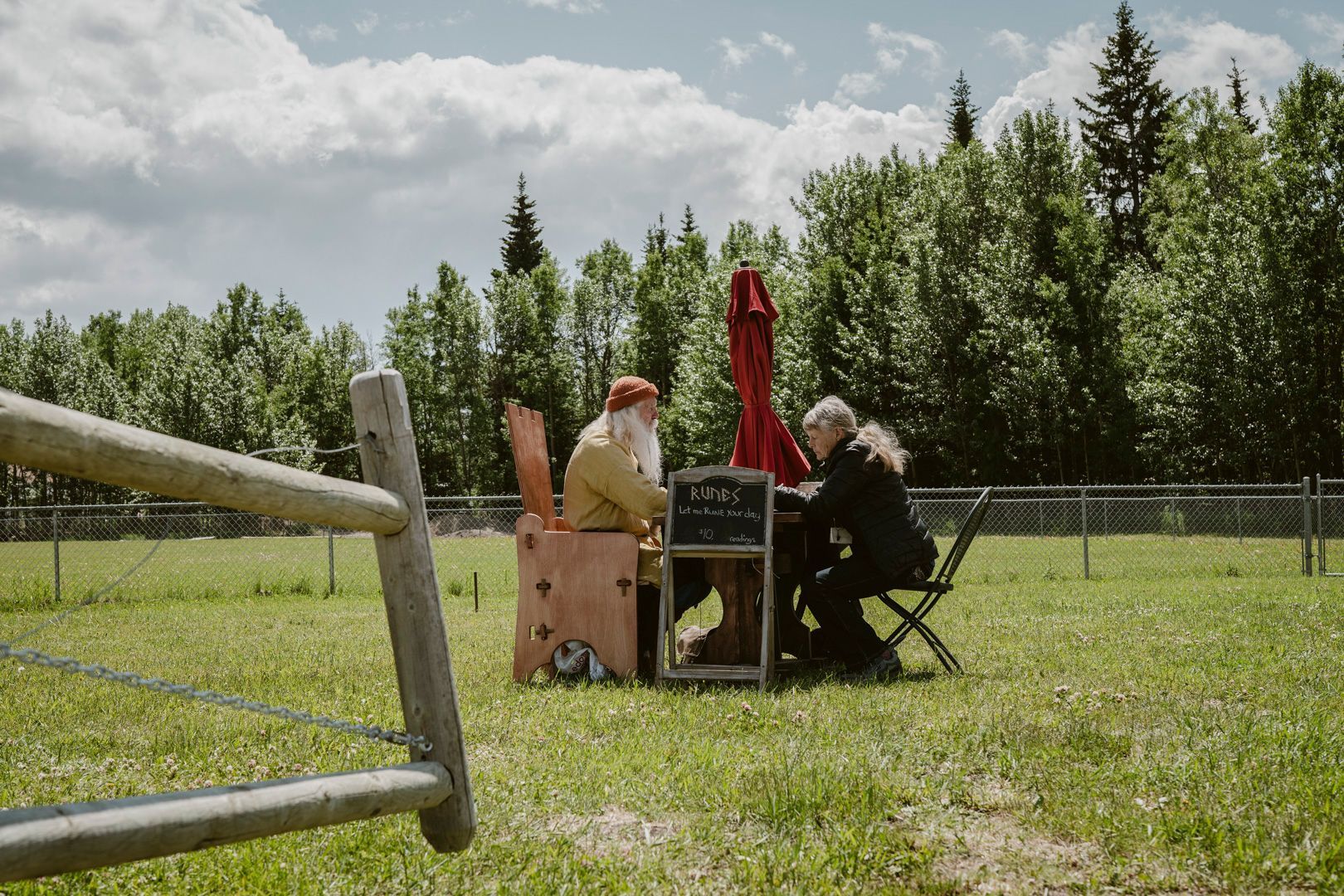 Two people sit at a table with a red umbrella in a field near a fence and a forest under a cloudy sky.