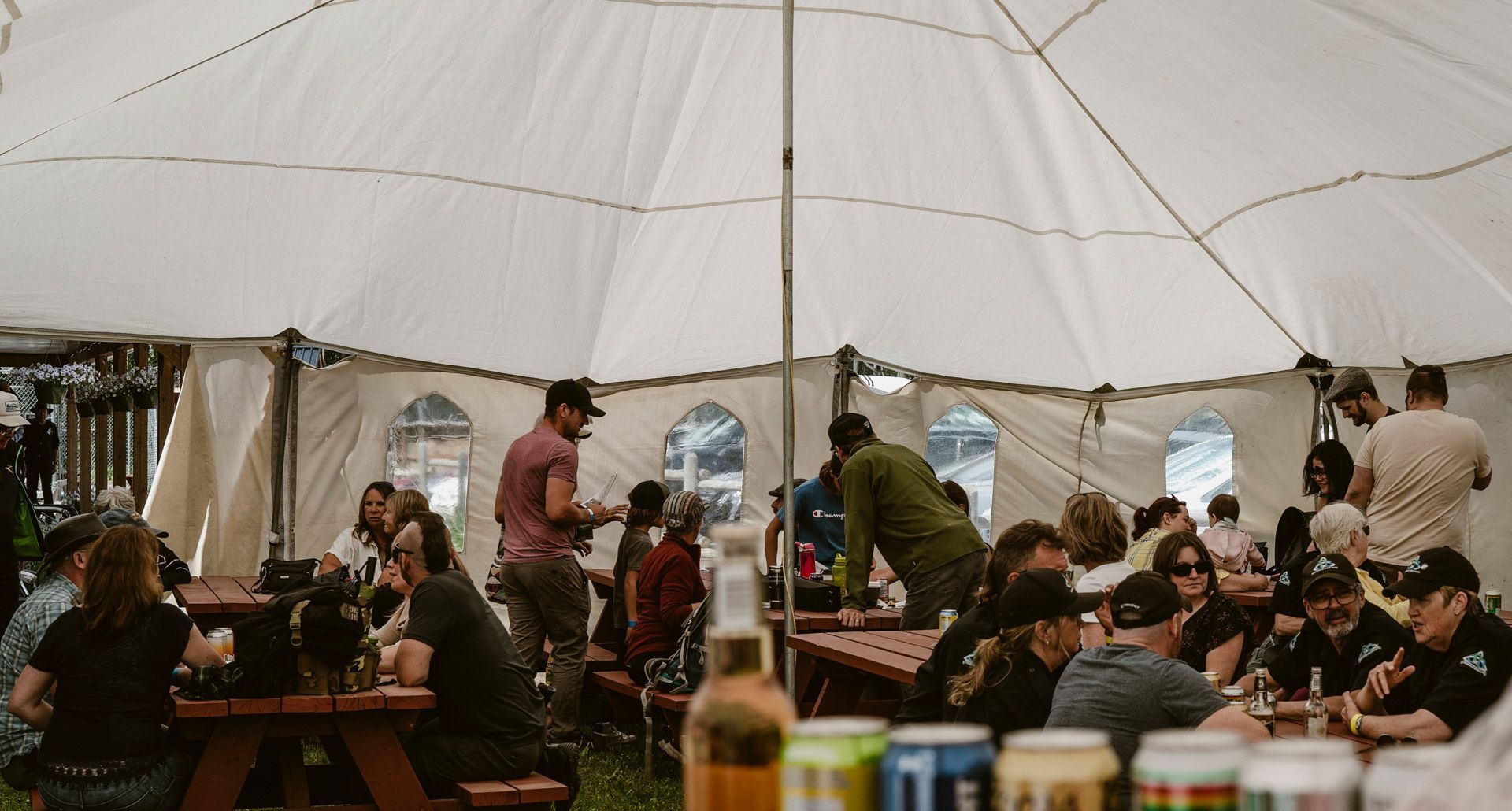 People gather at long tables inside a large, white event tent, drinking from cans and chatting.