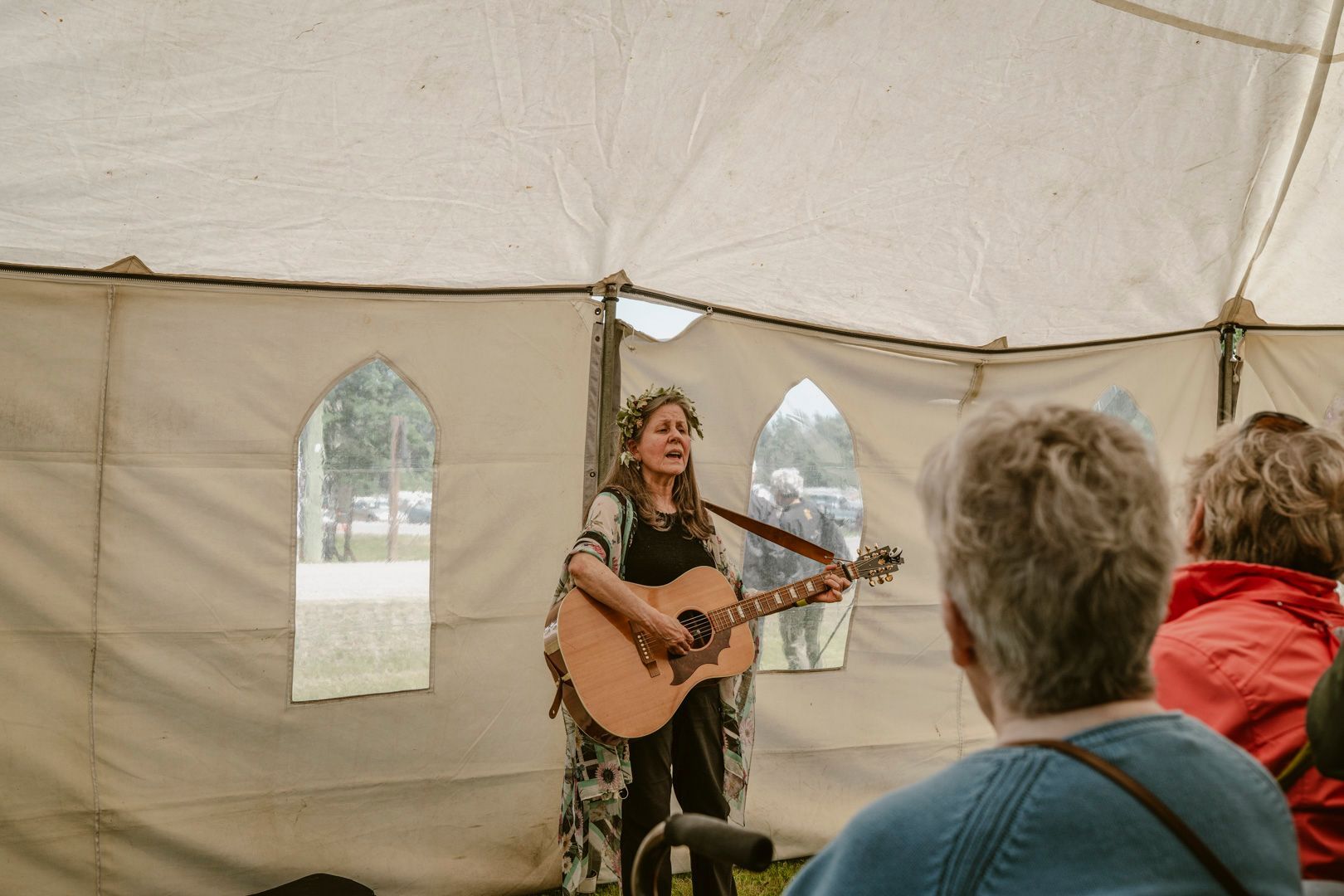 A performer wearing a floral crown plays an acoustic guitar inside a large, light-colored tent before an audience.