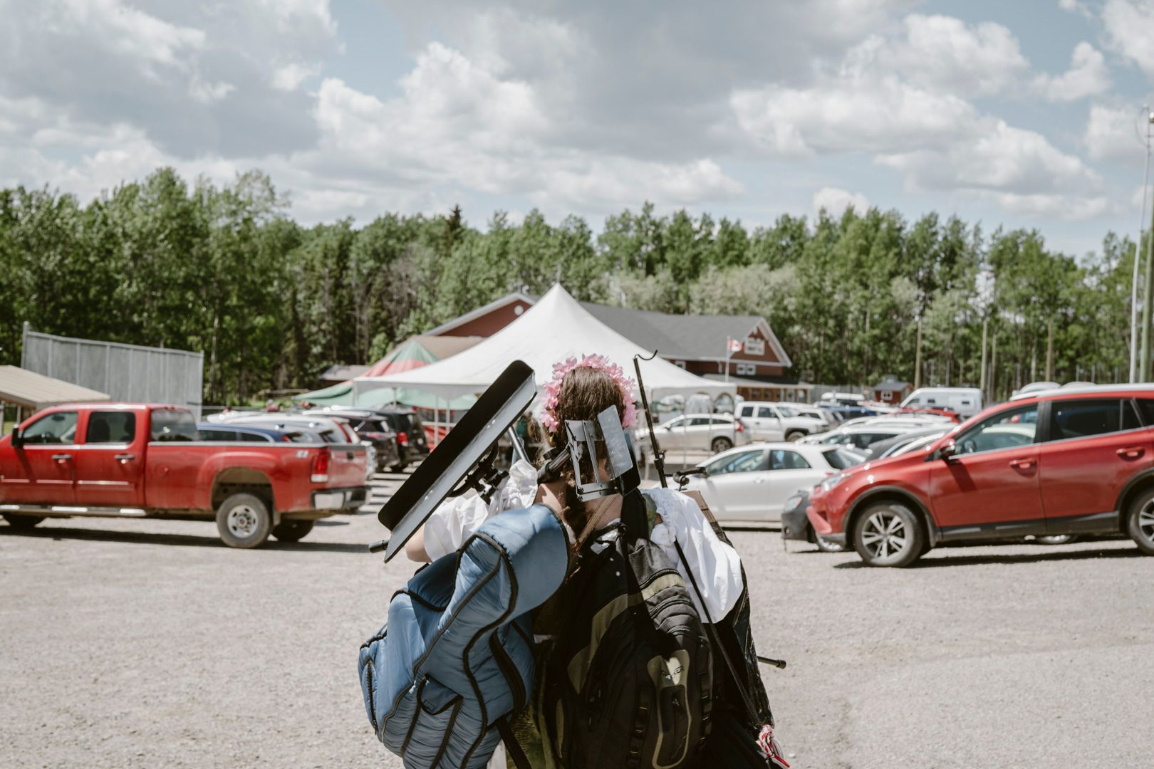 A person wearing a reflective silver mask and flower crown stands in a gravel parking lot with a blue musical instrument case.