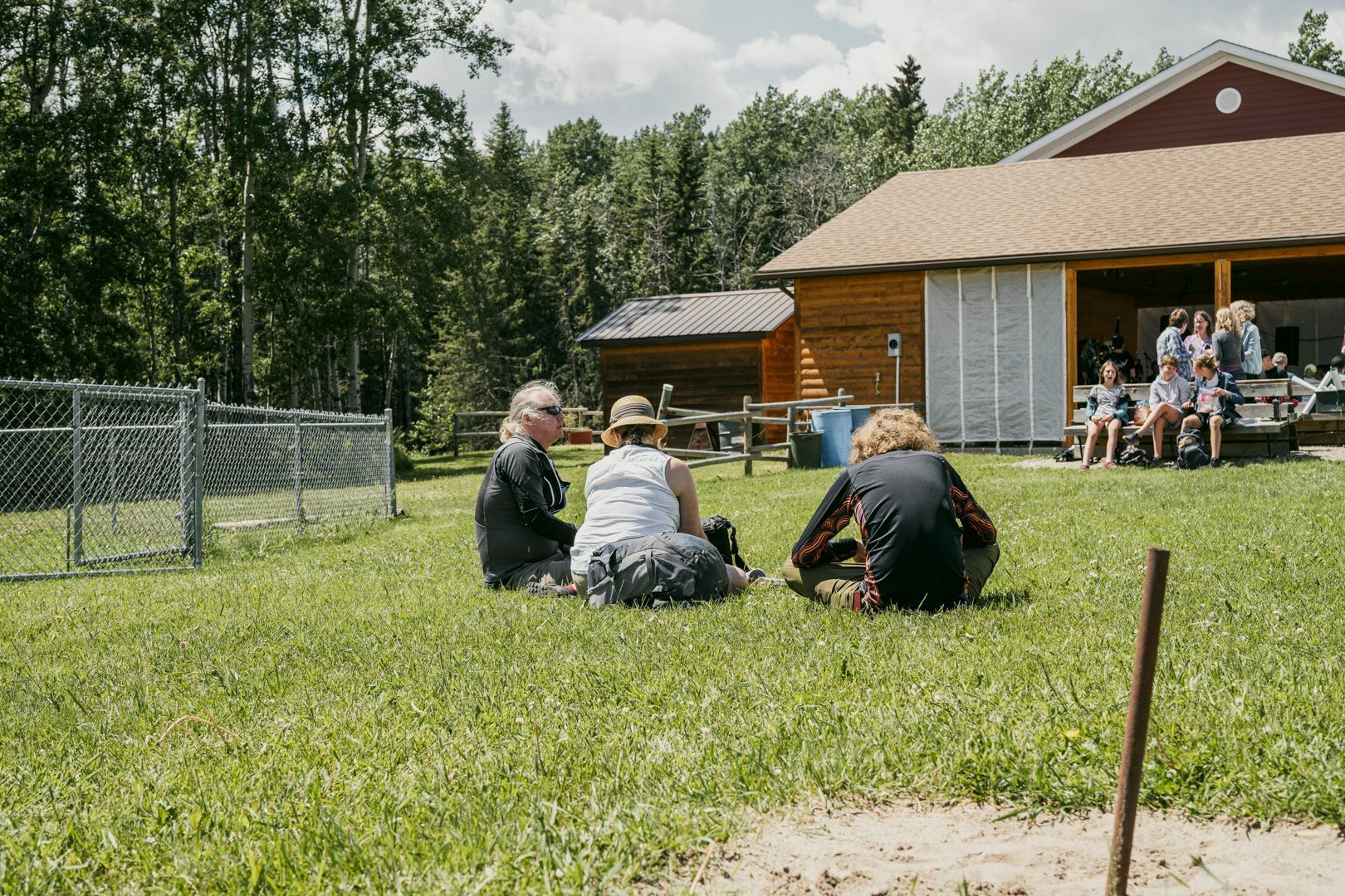 Three people sit on a grassy lawn facing a rustic wooden building with a covered porch near a wooded area.