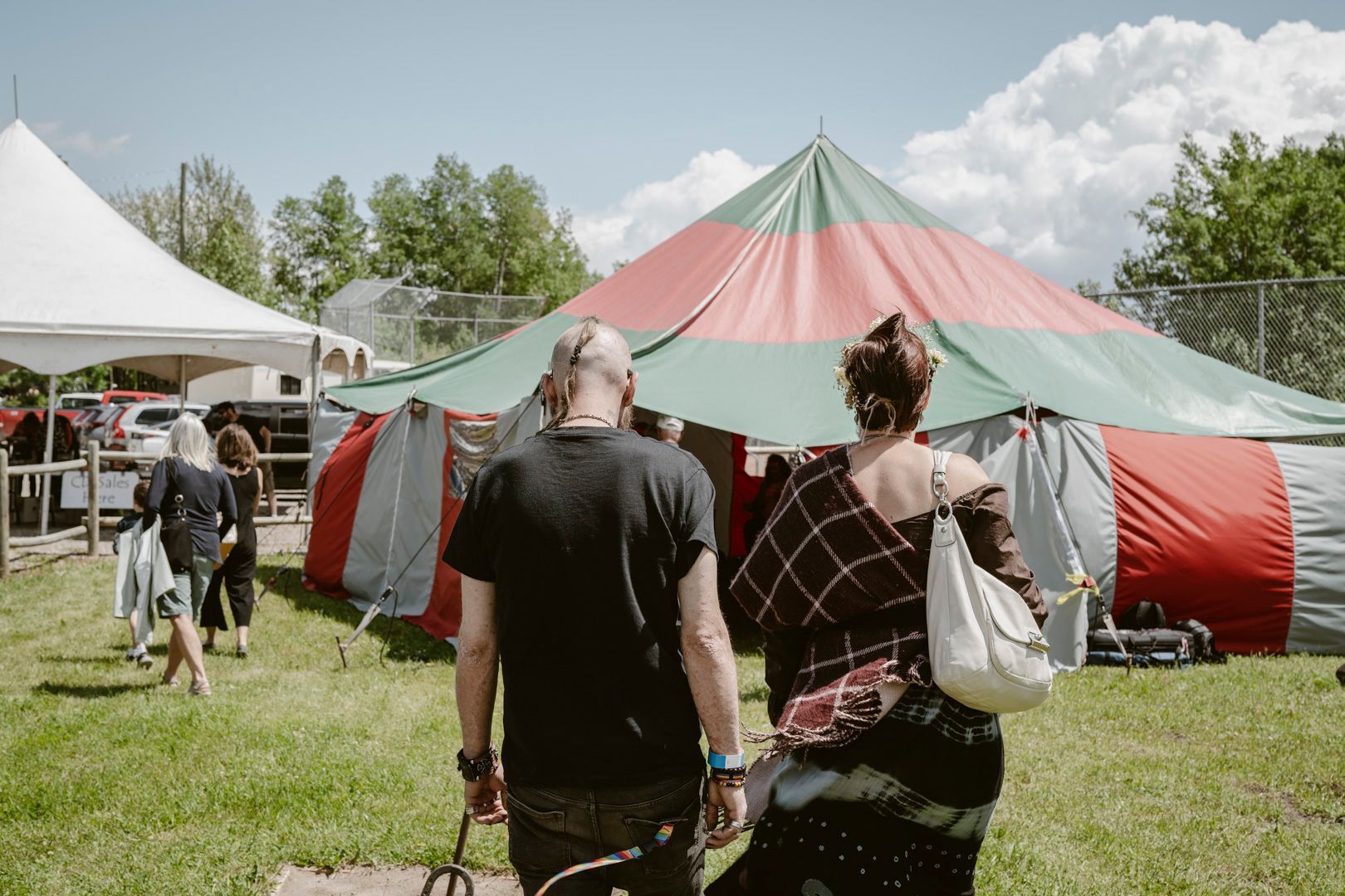 People walk toward a large red and green striped circus-style tent on a grassy field under a bright, sunny sky.