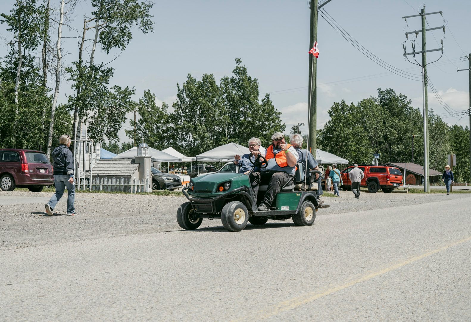 People ride in a green utility vehicle on a gravel road past a park with white tents and parked cars.