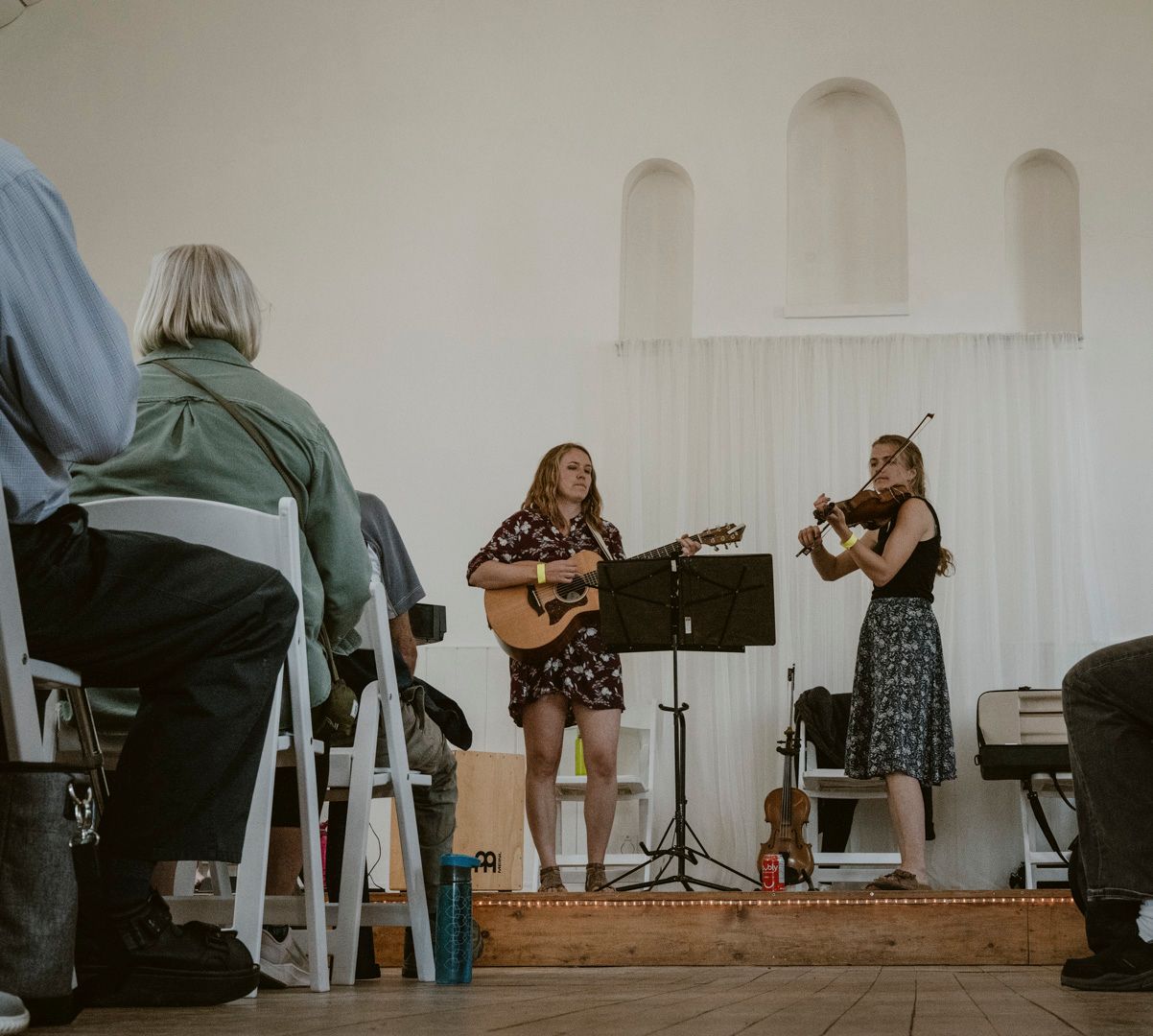 Two musicians perform on a stage in a hall; one plays an acoustic guitar while the other plays a violin.