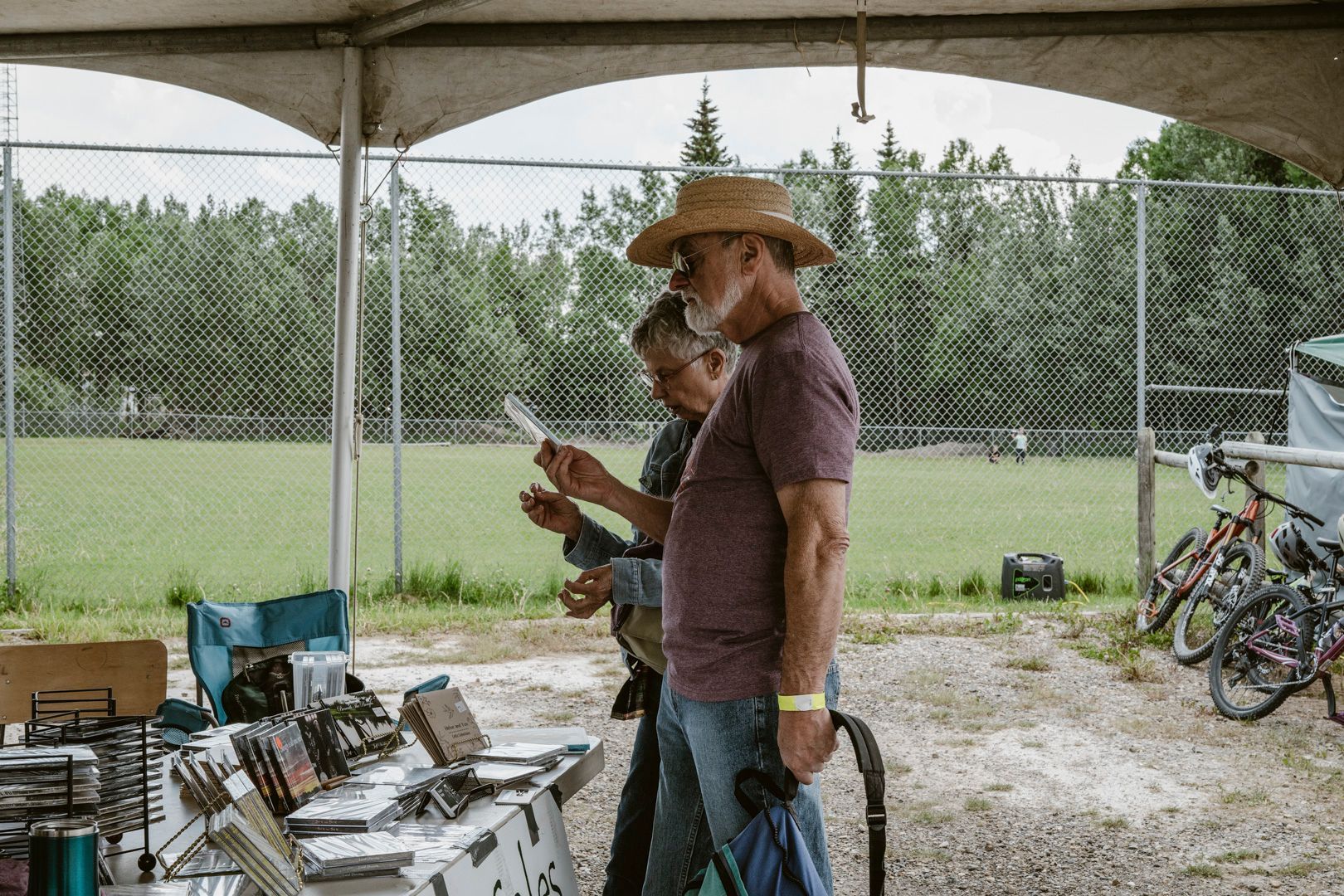 Two people in casual outdoor clothing stand under a canopy, examining small items at an outdoor stall near a fence.
