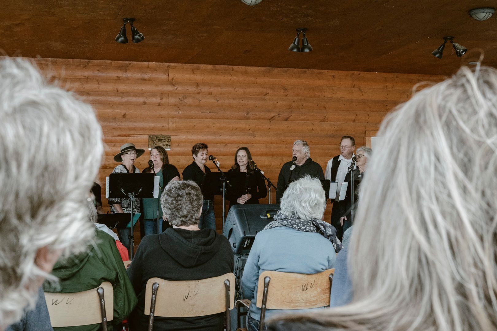 A group of people perform on a stage in a wooden building, viewed from behind an audience seated in chairs.