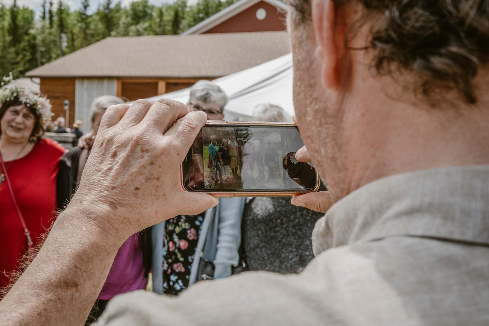 A person holds a phone to take a photo of a group of people standing outdoors near a red building and a tent.