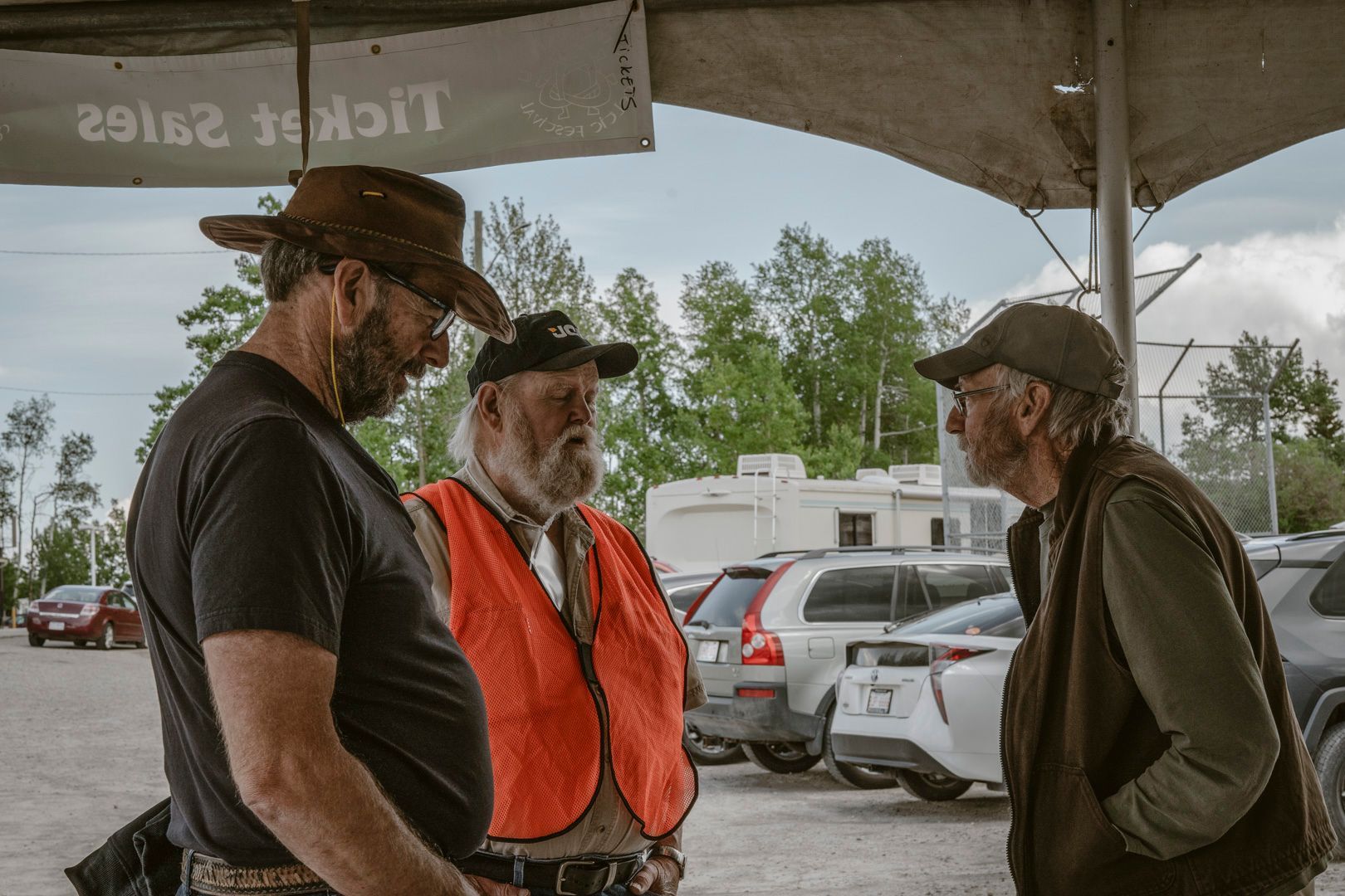 Three people in hats and outdoor clothing stand under a tent canopy, conversing in a parking lot.