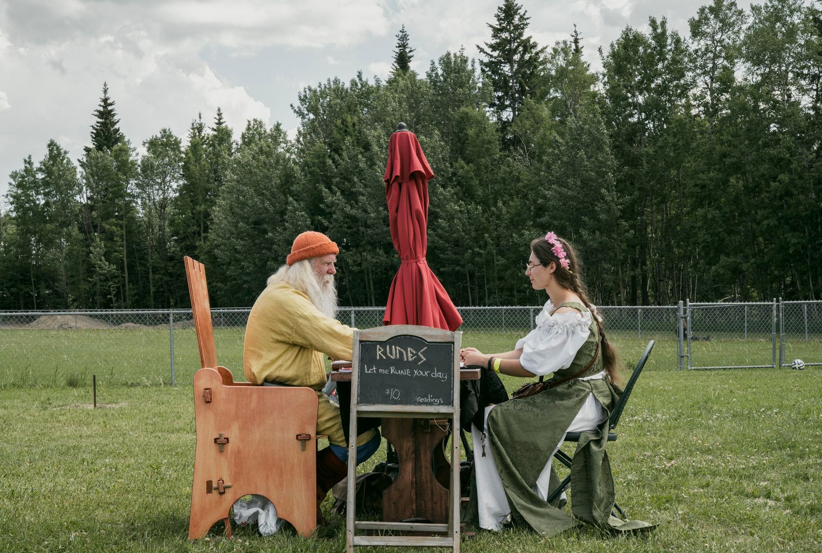Two people in medieval attire sit at a table outdoors, separated by a red umbrella and a small sign in an open field.
