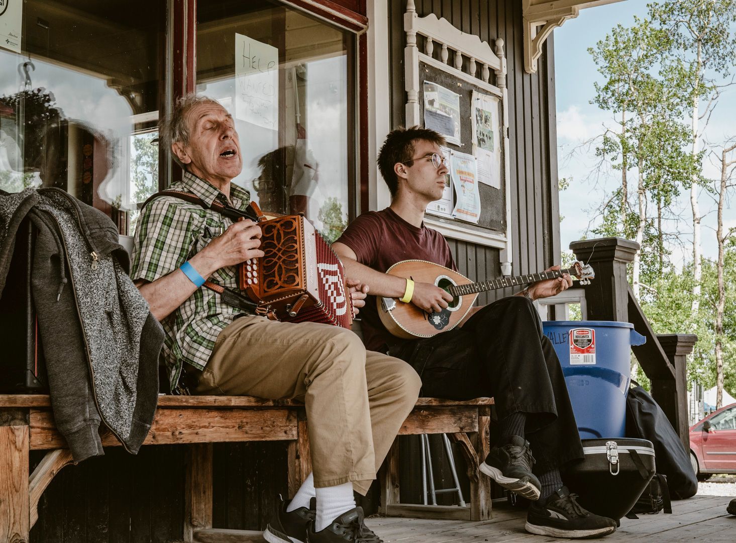 Two musicians play an accordion and a mandolin while sitting on a wooden porch outside a building.