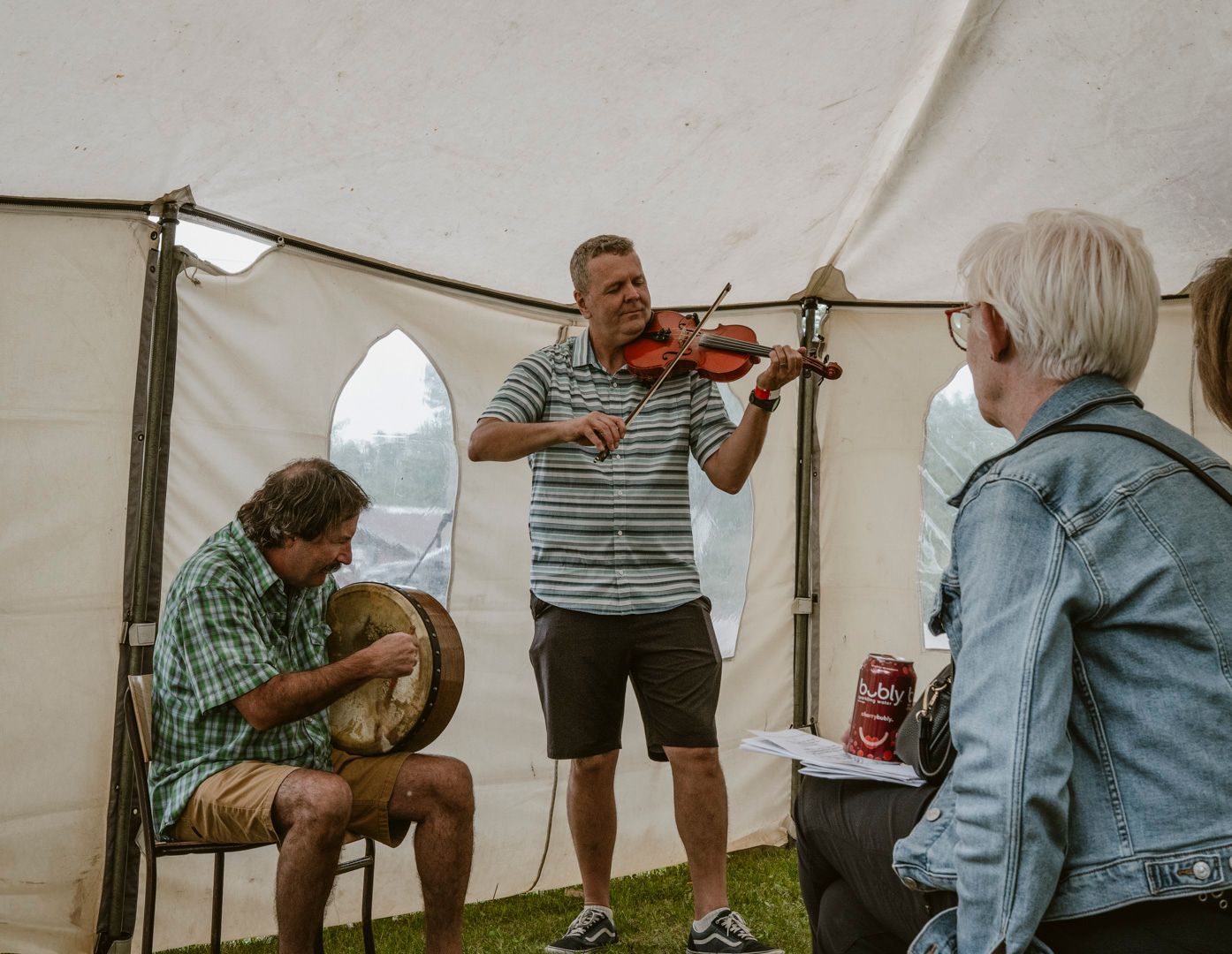 A musician plays the violin while another plays a hand drum inside a white event tent for an audience member.