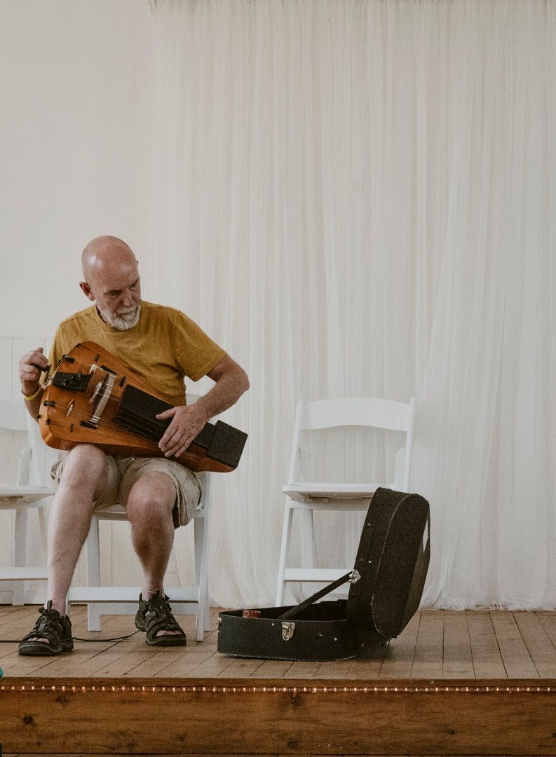 A person in a yellow shirt sits on a white chair playing a hurdy-gurdy, with an open instrument case on a wooden floor.