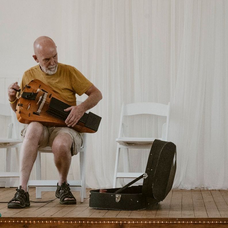 A person sits on a white chair on a wooden stage, tuning a hurdy-gurdy, with its open carrying case on the floor nearby.