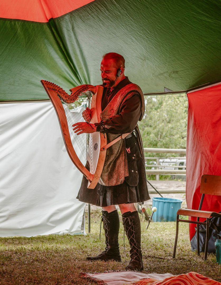 A performer in a kilt and tall boots plays a wooden harp while standing under a green and red tent.