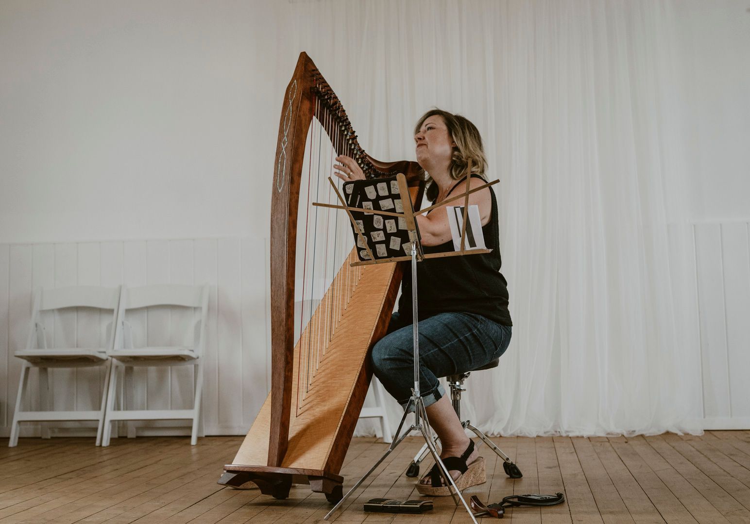 A musician plays a wooden harp while seated on a stool in a room with white curtains and two chairs.