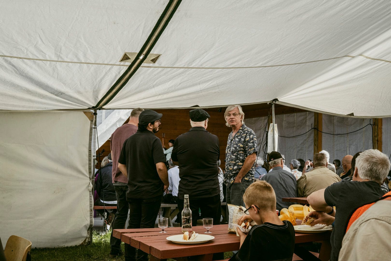 A group of people gather under a white canopy tent at outdoor picnic tables.