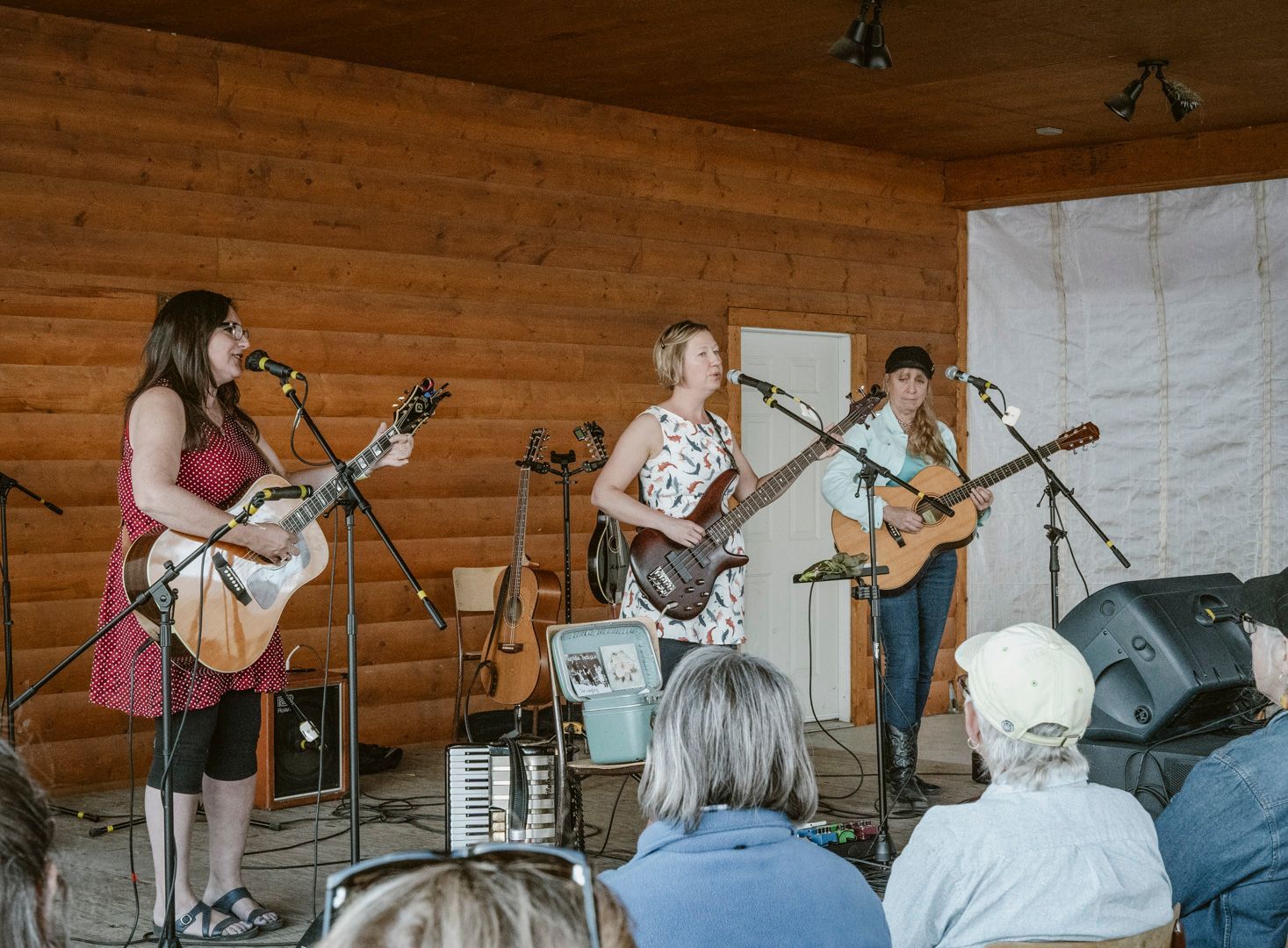 Three musicians play acoustic guitars and sing into microphones on a rustic stage before an audience.