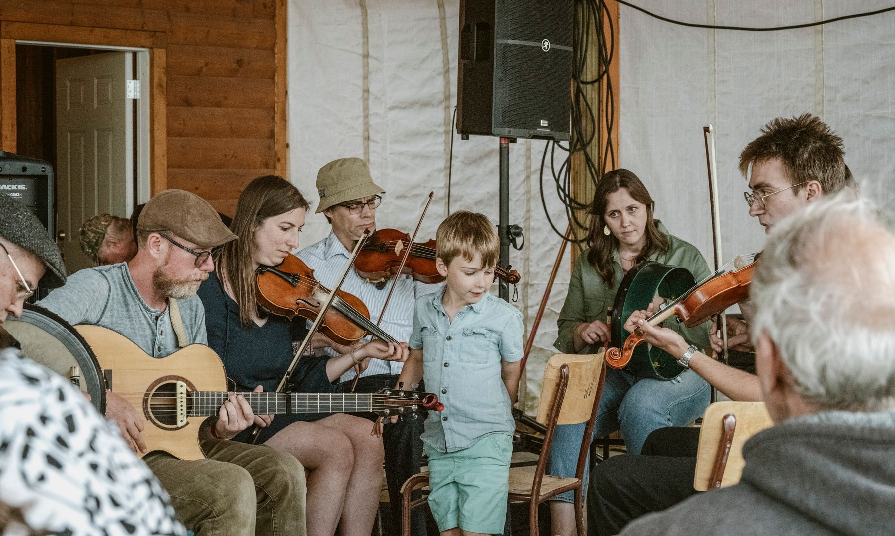 A group of musicians playing string instruments and acoustic guitar in an indoor setting with a child standing nearby.