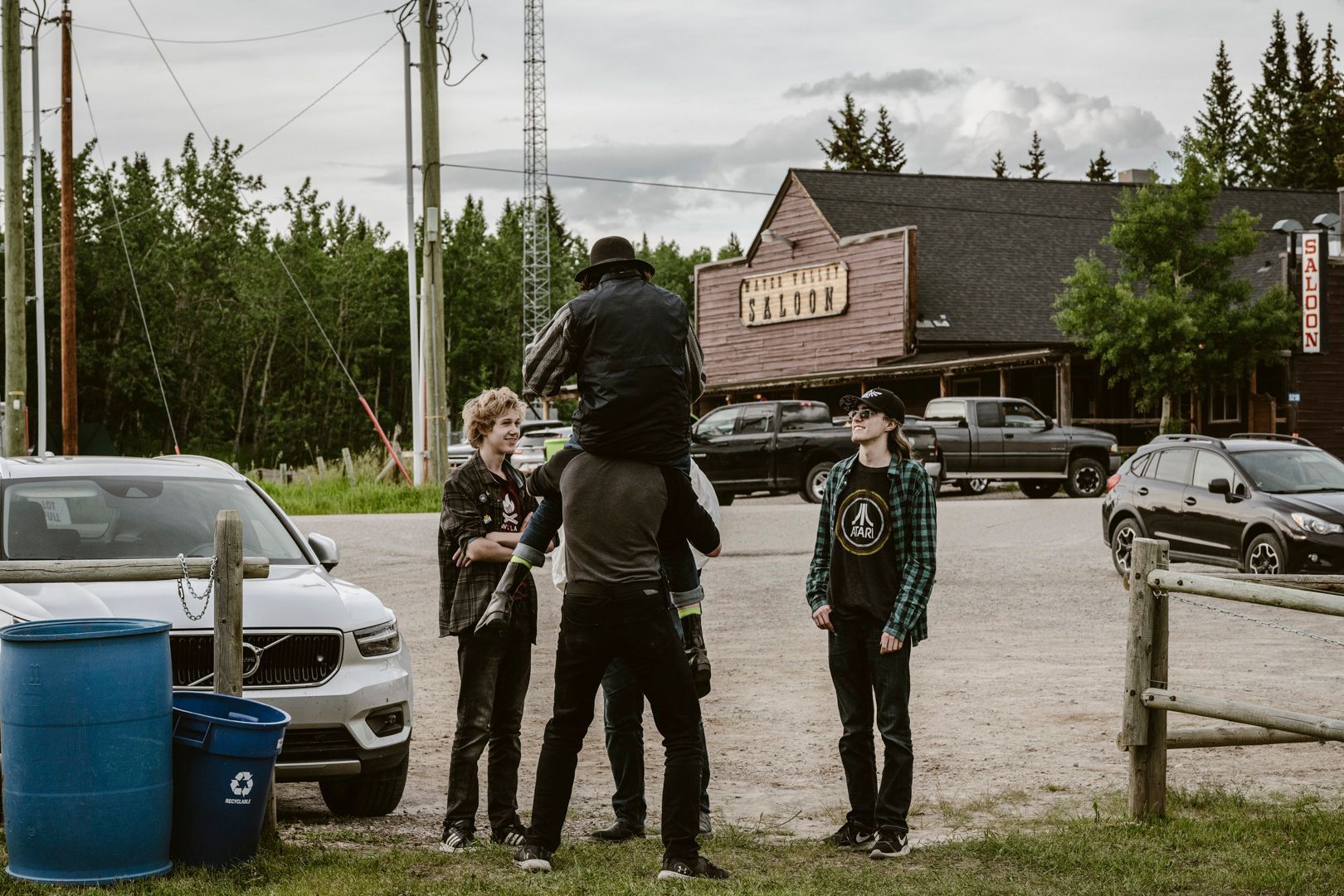 A group of people gathers in a gravel parking lot near a wooden building, with one person lifted onto another’s shoulders.
