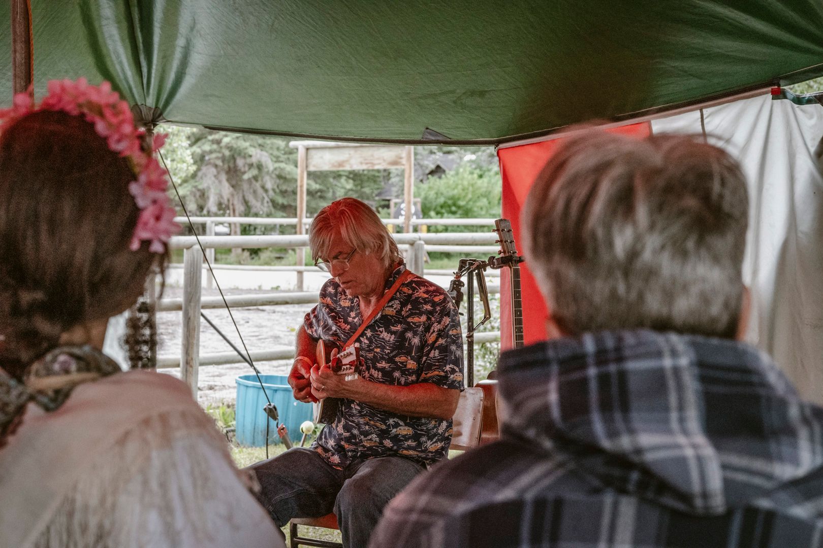 A performer plays a guitar under a tent canopy, seen from behind two audience members.