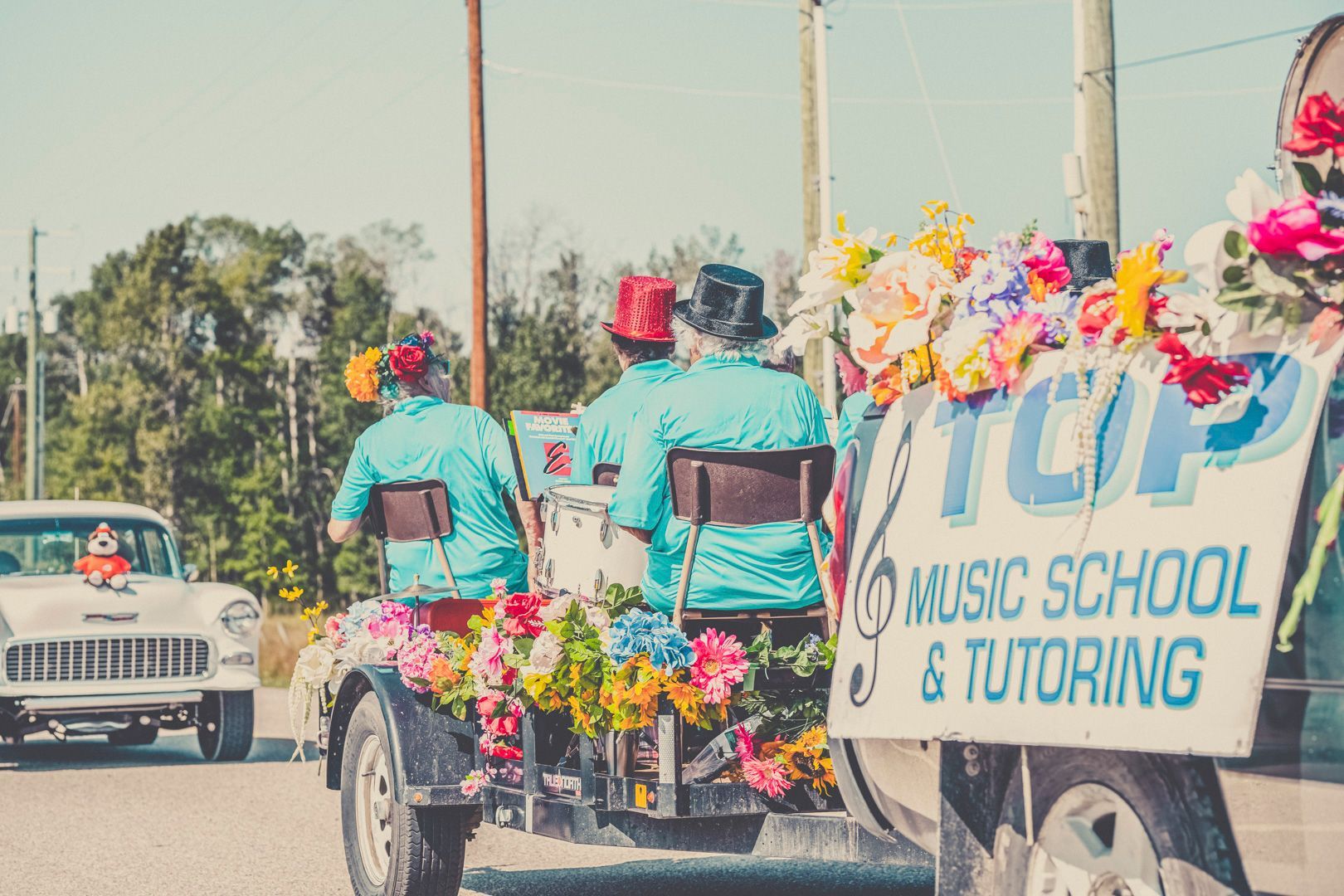 People in bright blue shirts and colorful hats ride a decorated trailer for a music school in a sunny parade.