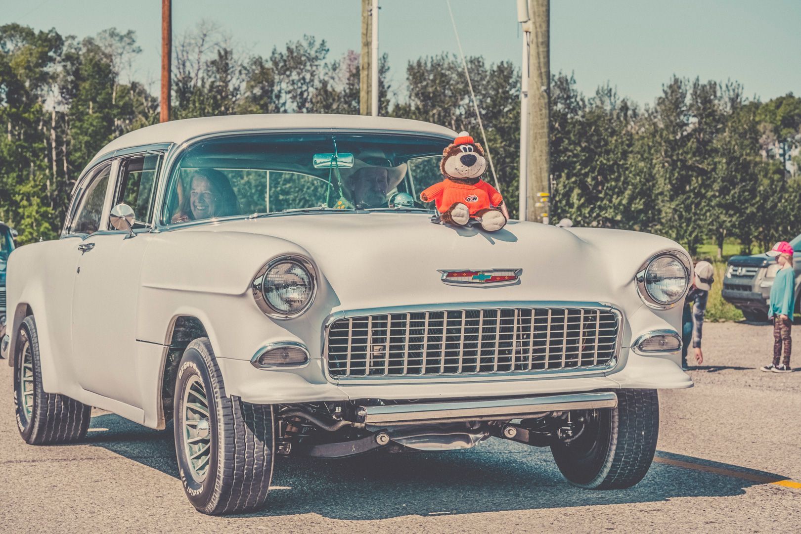 A white vintage Chevrolet car parked outdoors with a stuffed animal on its hood, with people standing nearby.