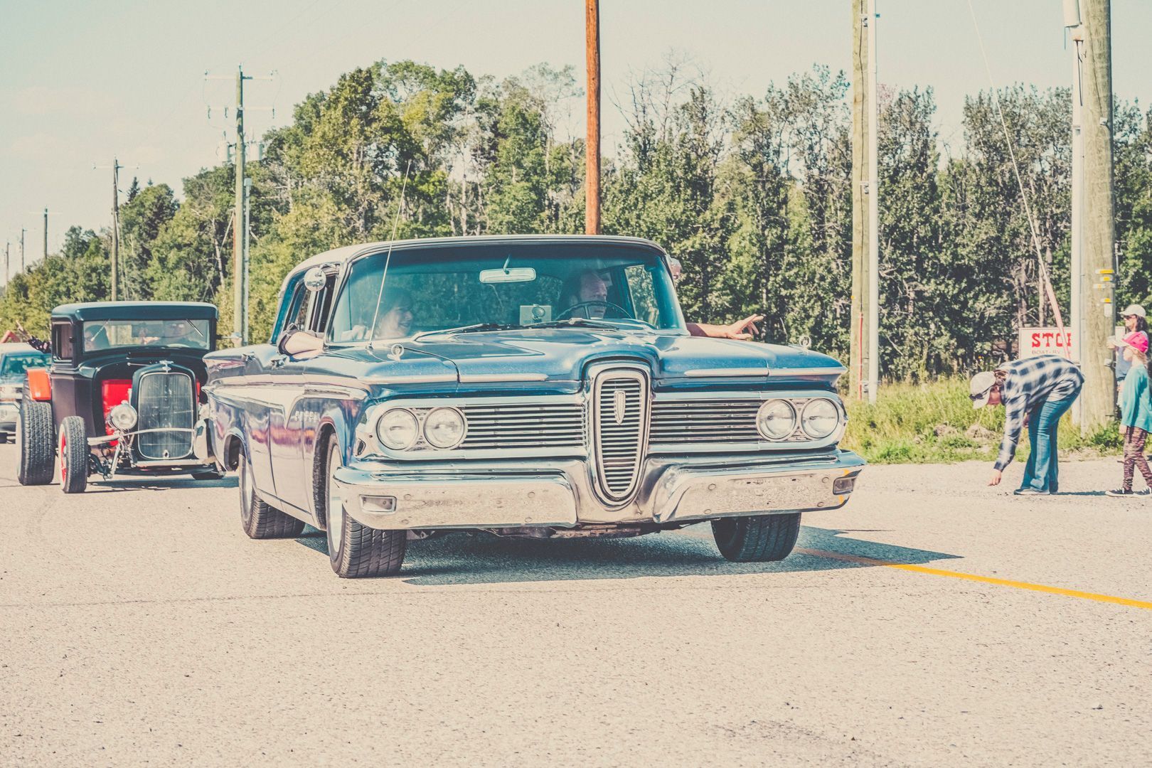 A vintage blue Edsel drives down a road, with a classic hot rod following behind and a person standing on the shoulder.