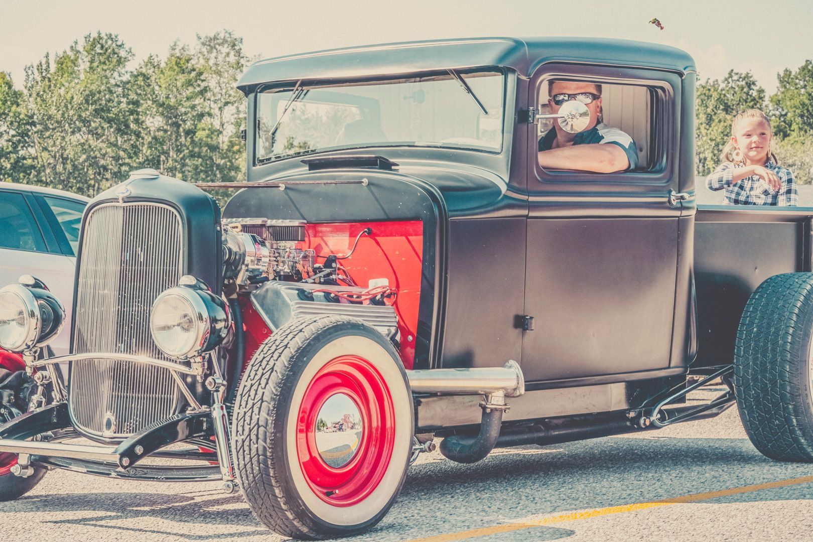 A black vintage hot rod truck with a red engine block and wheels, parked outside with a driver and passenger.