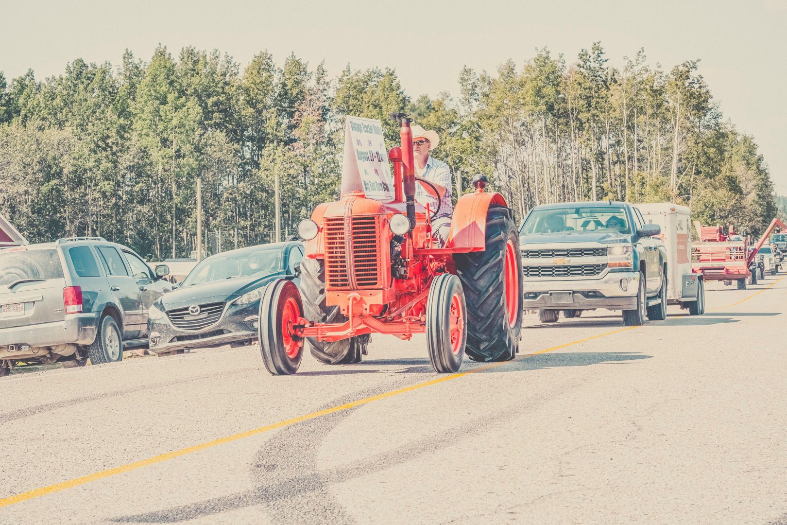 A bright red vintage tractor driving down a road in a parade with cars parked on the side.