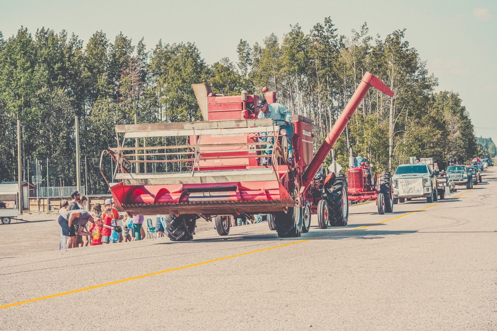 A large red combine harvester drives down a gravel road in a parade, followed by a line of vintage vehicles and tractors.