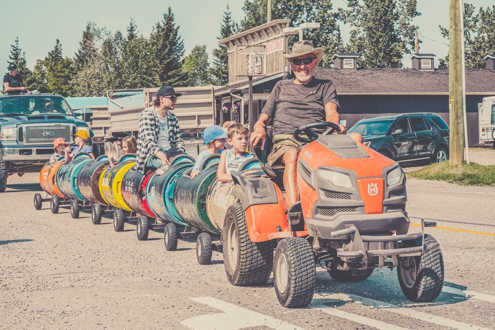 A man drives a riding lawnmower pulling a colorful train of barrel-shaped carts carrying children outdoors.
