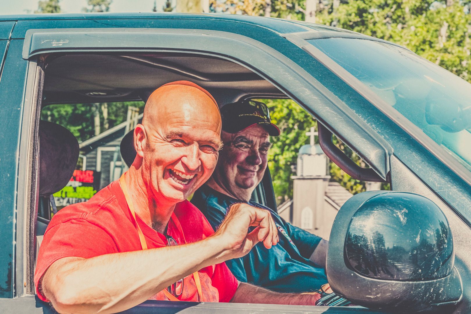 Two people smiling inside a vehicle; the driver in a red shirt gestures with his hand while the passenger wears a cap.