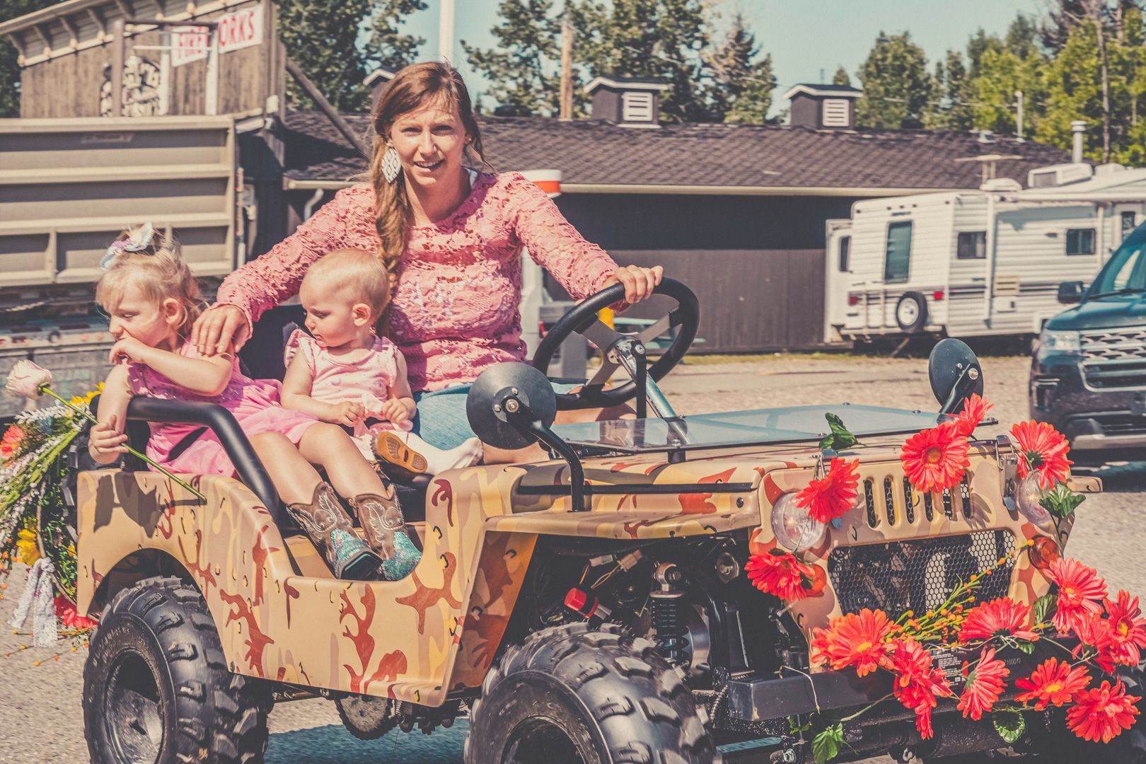 A person sits in a camouflage-patterned toy jeep decorated with red flowers, with two small children sitting beside them.