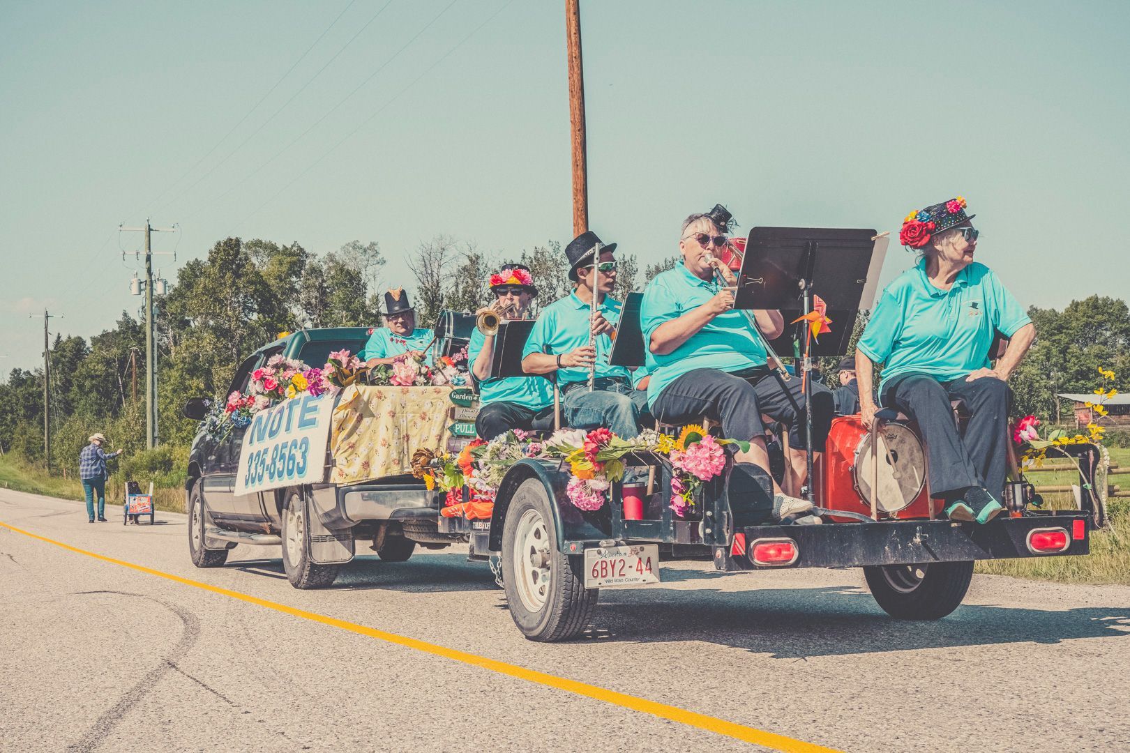 A musical group in teal shirts performs on a decorated flatbed trailer towed by a truck during an outdoor parade.