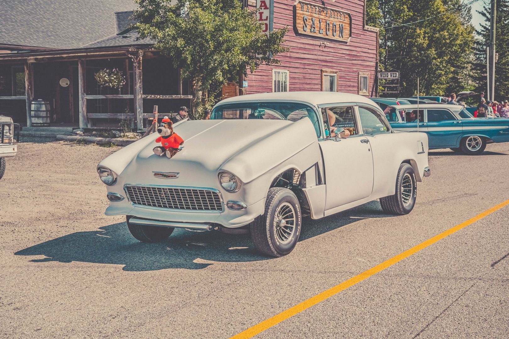A white classic car with its hood propped open sits in a gravel lot next to a wooden building, with a small dog on top.