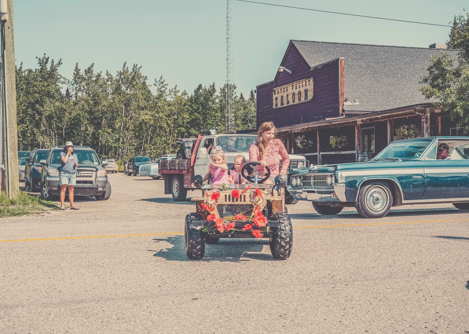 A decorated small, motorized toy tractor carrying two passengers drives through a parking lot in front of a rustic building.