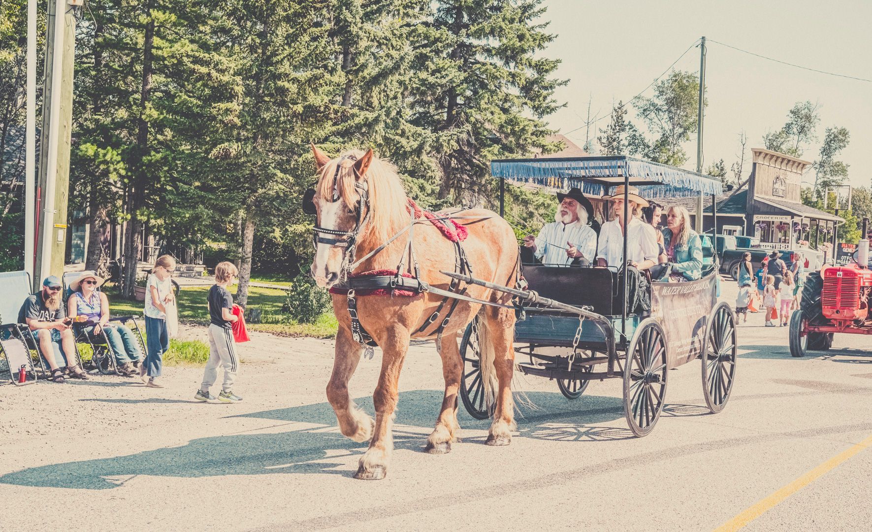A light brown horse pulls a carriage with passengers along a sunlit road in a small town, with spectators on the side.