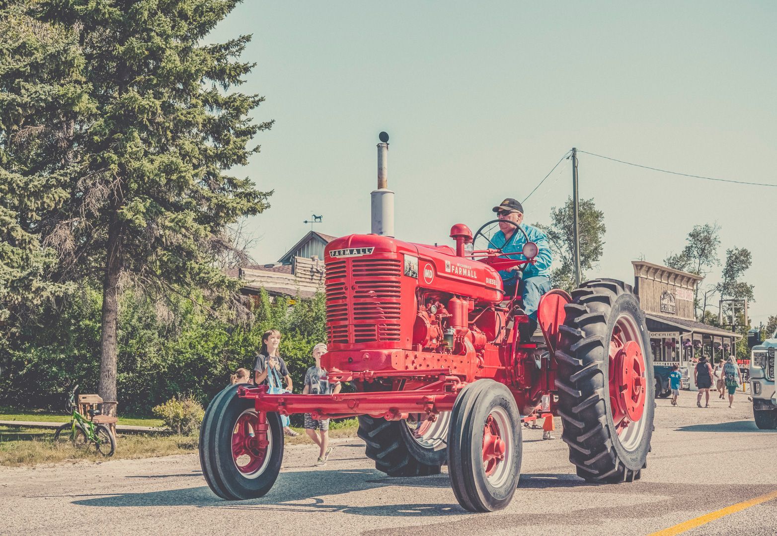 A man drives a vintage bright red tractor down a road past trees and buildings in a rural setting.