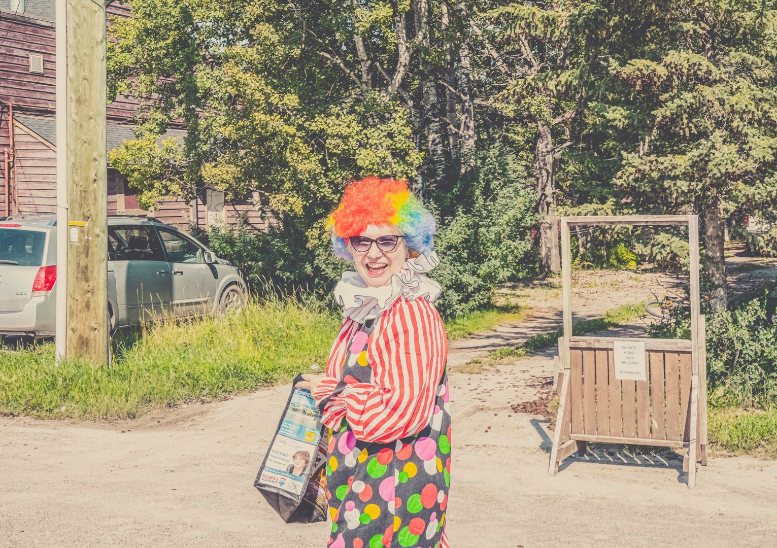 A person dressed as a clown with a colorful wig, striped shirt, and polka-dot pants smiles while standing outdoors.