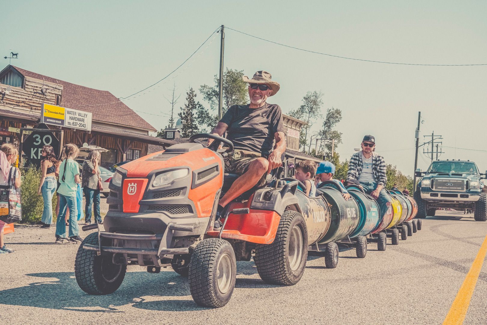 A person drives an orange lawn tractor pulling a train of colorful, barrel-shaped carriages along a sunny road.